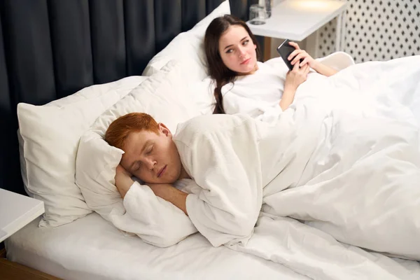 Красивая Молодая Женщина Лежит Постели Телефоном Смотрит Спящего Мужа — стоковое фото