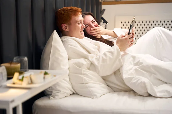 Смеющиеся Молодожены Отдыхают Гостиничном Номере Болтают Телефонам Большой Мягкой Кровати — стоковое фото