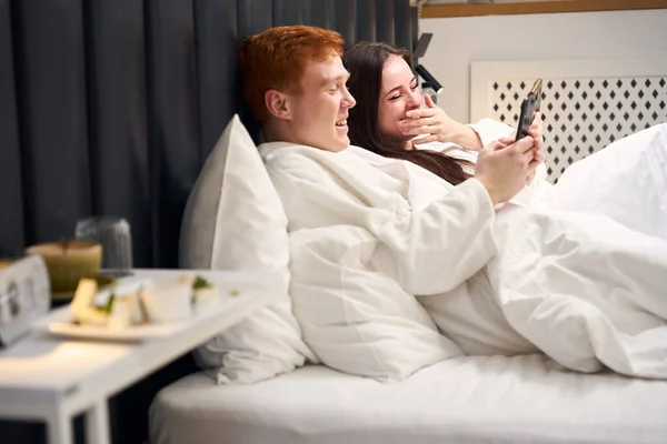 这对快乐的夫妇正在宾馆的房间里休息 在电话里聊天 他们躺在一张又大又软的床上 — 图库照片