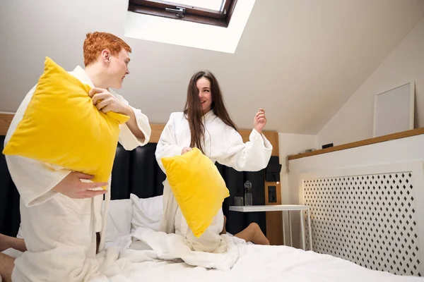 红头发的家伙和他的女朋友跳到床上和枕头打架 他们租了一间酒店房间 — 图库照片