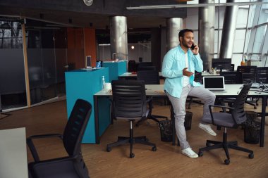 Masanın yanında dizüstü bilgisayarla duran ve iş yerinde telefonla konuşan bir erkek.