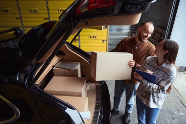 Arabanın bagajında elinde kutuyla duran ve kadına bakan gülümseyen genç bir adam.