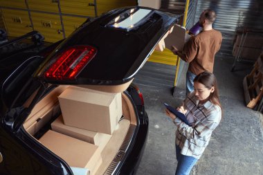 Genç bir kadın, kutularla dolu bir arabanın bagajında dururken deftere bir şeyler yazmakla meşgul.