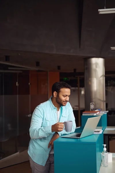 若い男がコンピューターの近くに立ってオフィスでカップを手に — ストック写真