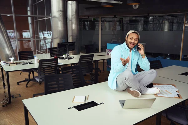 ノートパソコンの前の机の上に座ってオフィスのスマートフォンに電話している笑顔の男 — ストック写真