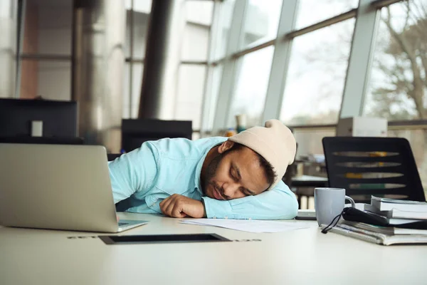 在商务中心 头戴头巾的年轻男性正在笔记本电脑旁的工作岗位上睡觉 — 图库照片