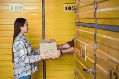 Depoda duran bir adama karton kutuyu gönderen gülümseyen kadının yan görüntüsü.