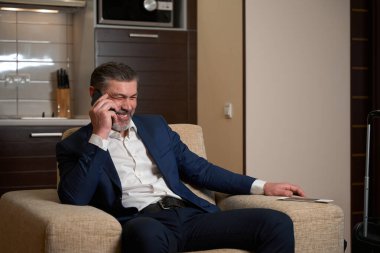 Ofis kıyafetleri içinde gülümseyen adam koltukta oturuyor ve pansiyonda akıllı telefon kullanıyor.