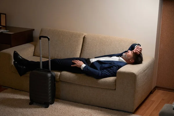 旅馆房间里 身穿办公室服装的成年男性躺在行李箱旁边的床上 — 图库照片
