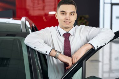 Genç adam ayakta duruyor, açık araba kapısına yaslanıyor, bir iş yeri yöneticisi bir otomobil filinde.