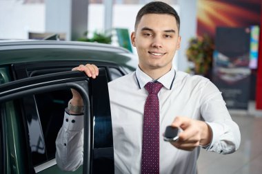 Çekici bir genç adam araba anahtarlarıyla açık bir arabanın yanında duruyor, günlük kıyafetlerle yönetici.