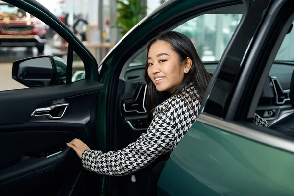 Happy young lady is sitting in the drives seat in a new car, the front door is open