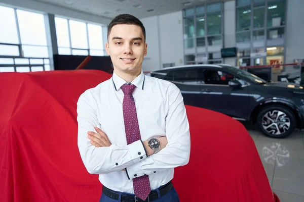 微笑着的年轻人站在一辆涂着红布的汽车前 经理正在经营着一家现代汽车经销商 — 图库照片