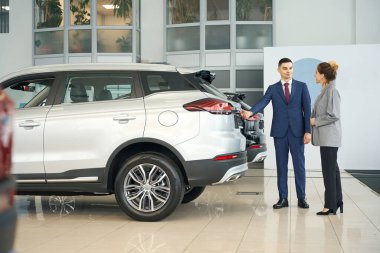 Takım elbiseli adam, araba galerisindeki mutlu kadına beyaz kesişme gösteriyor.
