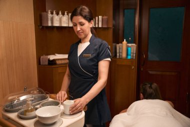 Yoğunlaştırılmış yetişkin ayurvedik uygulayıcısı seramik kaseye ilaçlı yağla dokunuyor.