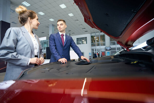 Smiling man and lady in suits standing near the hood of the car