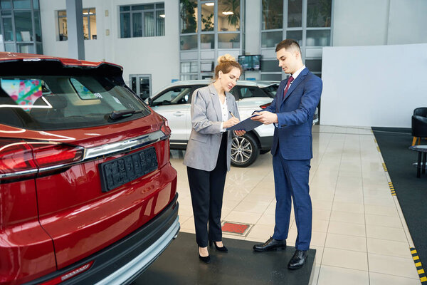 Lady and man in suits signing documents while standing near red automobile in car dealership