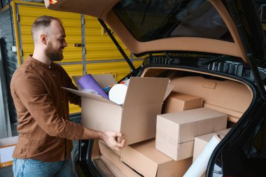 Tatmin olmuş bir erkeğin elinde bir kutu dolusu malla arabanın yanında duruşu.