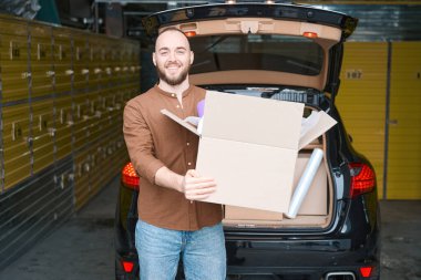 Elinde bir paketle depodaki arabanın yanında duran tatmin olmuş beyaz bir adam.