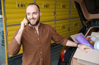Araba bagajının arkasında elinde malla gülümseyen bir erkek depoda akıllı telefondan konuşurken.