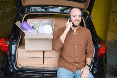 Memnun adam cep telefonuyla bir depodaki kutuların yanında sohbet ediyor.