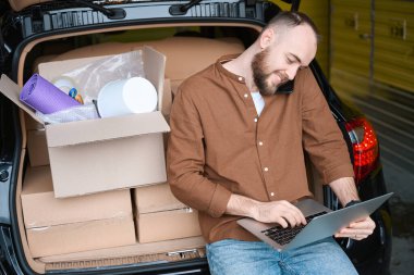 Mutlu yetişkin adam telefonda konuşurken dizüstü bilgisayarını kullanıyor. Arabasının yanındaki depoda duruyor.