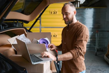 Depoda duran, kutularla dolu bir araba bagajında dizüstü bilgisayarını kullanan hoş bir genç erkek.