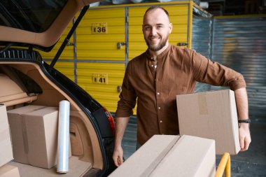 Canlı bir adam elinde bir kutuyla bir depoda elinde mallarla bir makinenin yanında.