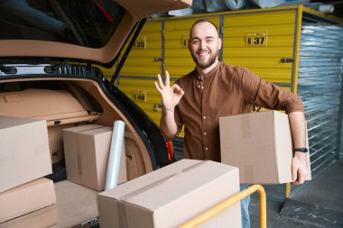 Elinde bir kutuyla gülümseyen erkek. Depodaki arabanın bagajının yanında.