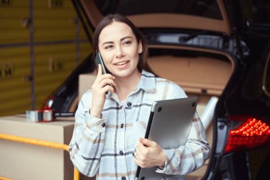 Elinde not defteri olan yakışıklı bayan akıllı telefonuyla konuşurken kutularla arabanın yanında duruyor.