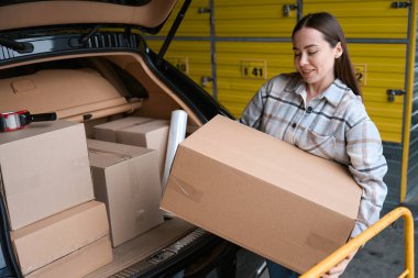 Gülümseyen genç bayan depodaki arabanın bagajında kutu koyuyor.