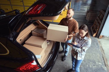 Odaklanmış bir adam bir arabanın bagajında kutu tutarken bir kadın bir şeyler yazıyor.