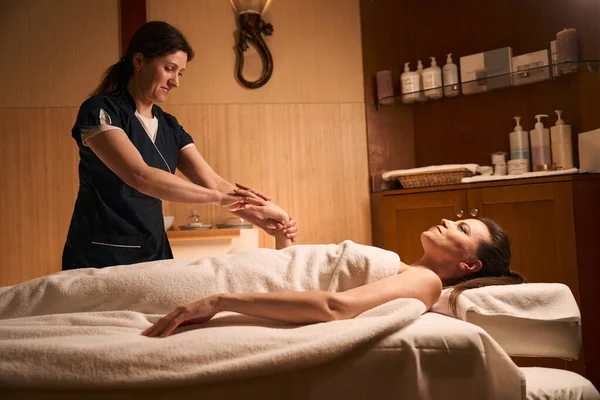 Smiling beauty clinic massotherapist stretching adult female patient fingers during massotherapy session