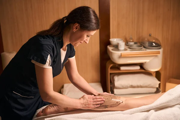 Focused massotherapist rubbing brown algae paste into skin on woman lower leg
