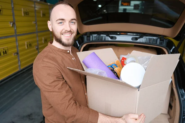 一个微笑的男人站在仓库里的汽车旁边 手里拿着一个盒子 — 图库照片