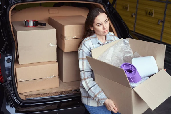 多愁善感的女人站在汽车行李箱旁边的仓库里 手里拿着装满商品的盒子 — 图库照片