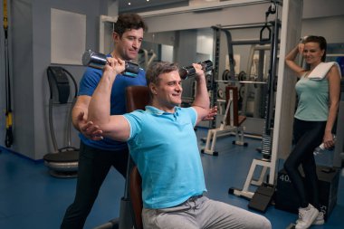 Keyifli sporcu, halterli omuz presini spor eğitmeni ve fitness partnerinin kontrolü altında yapıyor.