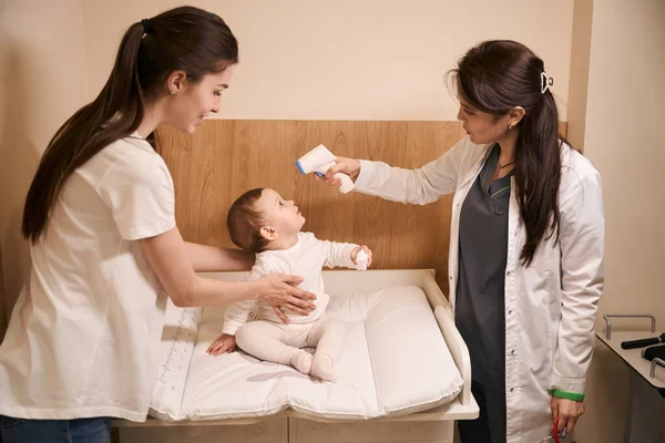 Poważny Pediatra Wskazujący Bezdotykowy Termometr Podczerwieni Czole Dziecka Obecności Uśmiechniętej — Zdjęcie stockowe
