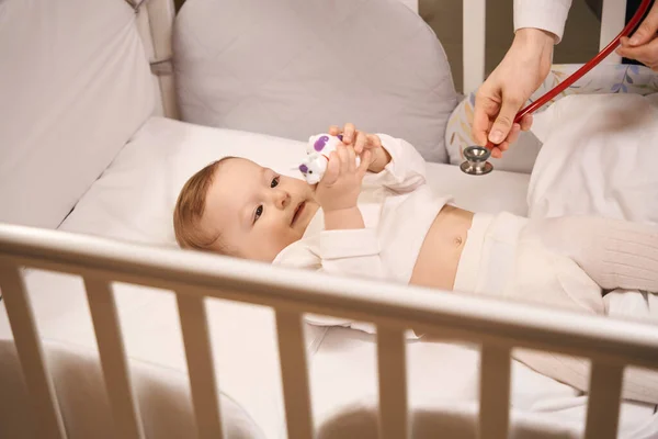 儿科医生 带听诊器的手伸向躺在婴儿床里抱着玩具的平静新生儿 — 图库照片