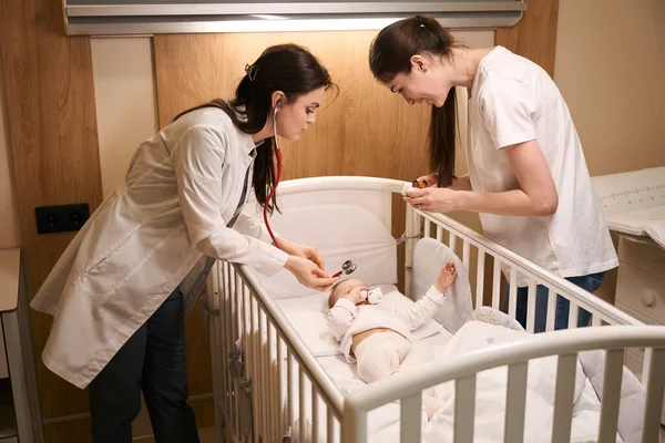 儿科医生在快乐的母亲面前用听诊器在婴儿床里给幼儿做检查 — 图库照片