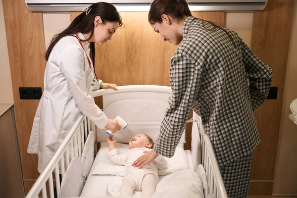 专注的儿科医生 当着高兴的女性家长的面 将非接触式红外温度计指向新生儿额头 — 图库照片