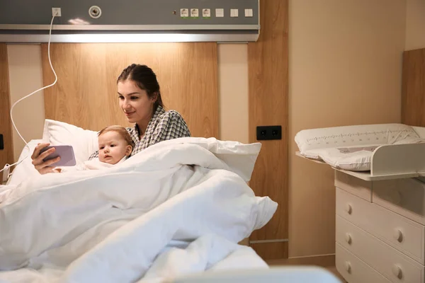 快乐的年轻妈妈用智能手机向躺在病床上的生病的孩子演示视频 — 图库照片