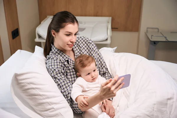 女家长在手机上向躺在病房里的婴儿展示卡通片 — 图库照片