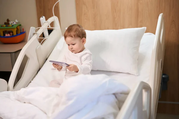 在医院病房里 手里拿着手机的病童一个人坐在床上 — 图库照片