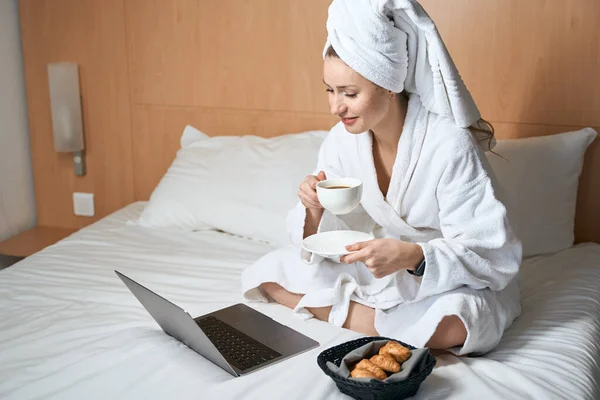 ホテルの部屋でベッドの上にクロワッサンとノートパソコンのコーヒーを飲むバスローブの女性の写真 — ストック写真