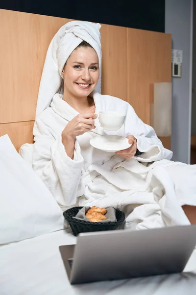 バスローブにコーヒーと幸せな女性の垂直写真 コーヒーを飲むクロワッサンとベッドの上にノートパソコンに座って — ストック写真