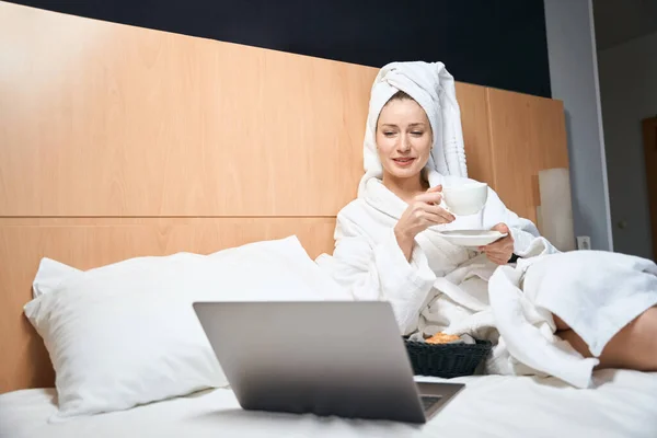 白いバスローブに身を包んだ女性の写真付きのラップトップは ホテルの部屋でベッドの上にコーヒーと座って — ストック写真
