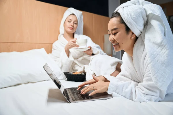 ホテルにはノートパソコンとコーヒーを備えたガールフレンドがリラックスしています 二人の女性が白いコートでベッドの上で話している — ストック写真