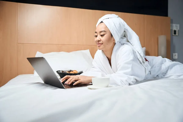 ホテルのベッドでラップトップの近くのバスローブのアジアの女性 ホテルの部屋でコーヒーやクロワッサンとリラックスした女性 — ストック写真