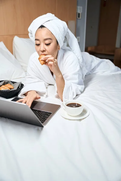 ノートパソコンでベッドでクロワッサンをかむバスローブの魅力的な女性 白いバスローブを着た女性がホテルの部屋で休んでいる — ストック写真